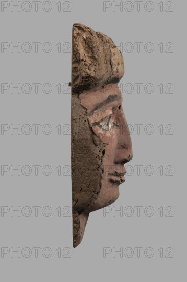 Sarcophagus mask with a partially preserved fragment of a wig above the forehead painted yellow with traces of black polychrome. It is separated from the forehead by cherry-red and black bands. The eyes are relatively large, with black rims, pupils, and eyebrows; the whites are white. The nose is long, quite narrow, widening evenly towards the nostrils. The mouth is primitively modeled. A fragment of the neck is preserved under the chin. The face is painted cherry-red. Partially preserved mounting pegs at the back. Ptolemaic Period, 332–30 BC, Egypt. Sarcophagus mask; height 25.7 cm, width 18 cm, thickness 8.2 cm., A side view of a carved face fragment made of rough, textured material in shades of brown and pink, showing a pronounced nose, lips, and a stylized eyebrow and eye area with traces of white and darker pigment, set against a flat gray background.