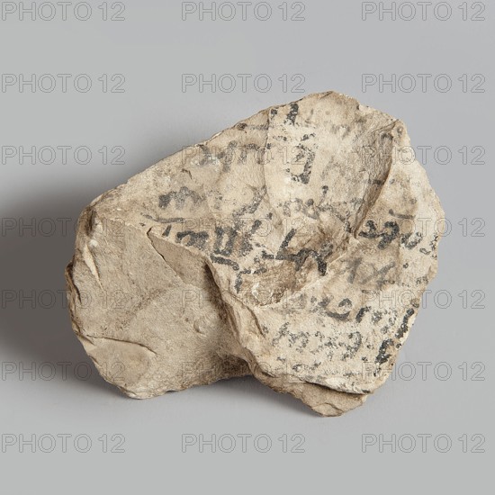 Ostracon with lines of Coptic text — 7 on one side, 2 on the other; Coptic ostracon; unknown creator; ; 7th–8th century, Egypt; ostracon; height 9.7 cm, width 7.5 cm, depth 2.3 cm., A roughly triangular, irregularly shaped piece of light beige stone with a textured, pitted surface sits against a plain light gray background, showing several darker gray and black smudges and markings across its upper face, along with areas of shadowed recesses and a few smoother, flatter planes around its edges; the stone casts a soft shadow on the background and displays subtle variations of tan, cream, beige, light brown, gray, and black tones.