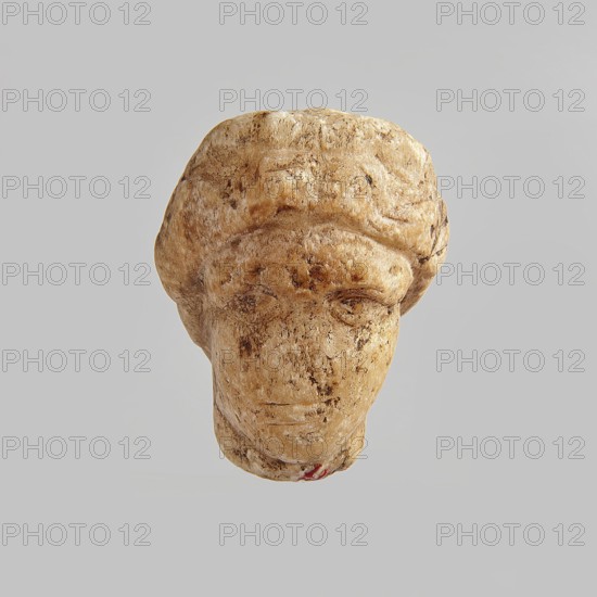 Head of a woman, Roman period, Egypt; figurine; height 5.5 cm, width 3.5 cm, depth 4.5 cm., A carved stone head with worn facial features and a rounded hair-like shape, showing a weathered surface with rough texture and small pits; the object appears in shades of beige, tan, brown, and cream with darker brown speckles and smudges across the face and hair area, and a small touch of red near the bottom; the background is a uniform light gray.