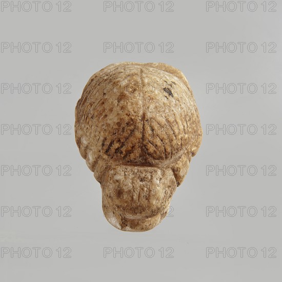 Head of a woman, Roman period, Egypt; figurine; height 5.5 cm, width 3.5 cm, depth 4.5 cm., A roughly textured object with a rounded top and a protruding lower section, appearing carved from a light brown stone with darker brown mottling and areas of white. The surface shows carved grooves and ridges forming symmetrical patterns on the rounded top and sides, with visible wear and small dark spots. The background is a smooth, pale gray.