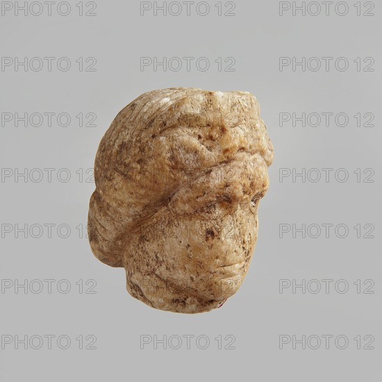 Head of a woman, Roman period, Egypt; figurine; height 5.5 cm, width 3.5 cm, depth 4.5 cm; creator, ., A worn stone sculpture of a human head shown against a plain light gray background, with a rounded hairstyle swept back and a smooth, weathered surface that shows patches of darker brown and beige tones, softer cream and tan areas, and some darker speckling; facial features are smoothed but include a brow, eye contours, a nose, and a mouth, with overall natural stone textures and subtle grooves.