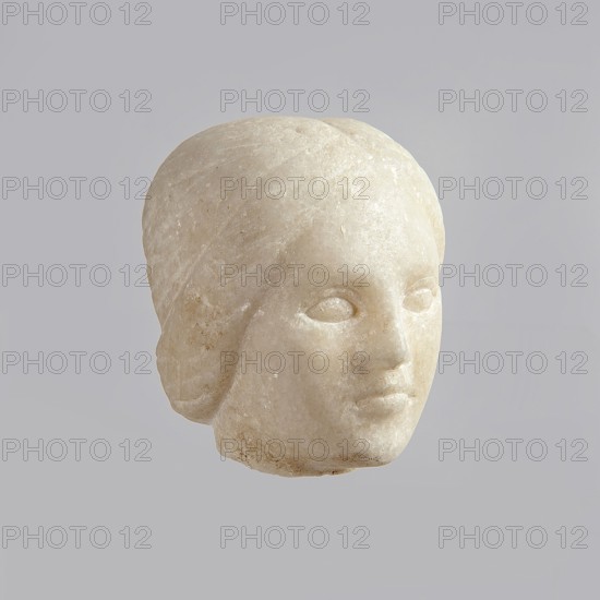 Fragment of a female figurine. Fragment of a small-sized sculpture or sculptural group. The depiction has portrait features. The face is oval, the nose is large, the mouth is closed in a slight grimace. 2nd–3rd century AD, Egypt. Figurine, sculpture; height 8.5 cm, width 4.5 cm, depth 5.5 cm., A pale stone sculpted head with smooth rounded contours, showing a stylized face with closed eye sockets, a defined nose and lips, and hair shaped close to the head; the stone is off-white to cream with subtle darker beige speckles and slight surface weathering, set against a uniform light gray background.