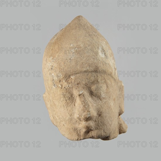 Head of a man, Archaic Period, Cyprus; figurine; height 7 cm, width 5 cm, depth 5.5 cm., A worn sculpted head in beige and light brown tones set against a pale gray background, showing a pointed top and rounded facial features with a smooth yet pitted surface, faintly defined eye sockets, a broad nose, subtle mouth, and a few rough textured areas and small darker speckles across the surface.