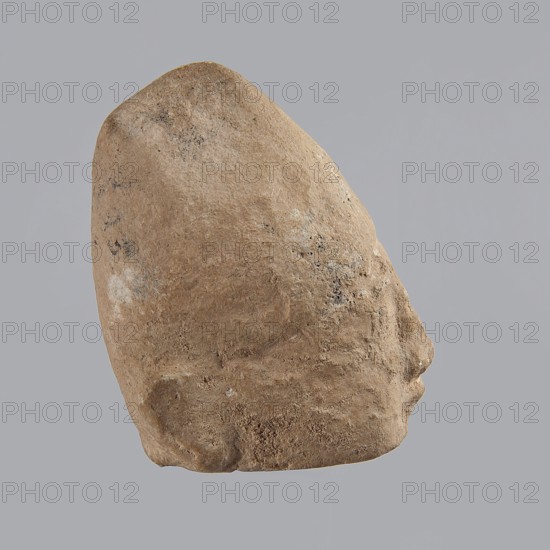 Head of a man, Archaic Period, Cyprus; figurine; height 7 cm, width 5 cm, depth 5.5 cm., A weathered, irregularly shaped stone object with a rough, textured surface in shades of tan and light brown, with small patches of darker brown and faint areas of off-white; the object is shown in profile against a flat, pale gray background, revealing a rounded top, a sloping side, and a subtle protrusion near one end, with varied surface pitting and scattered darker speckles.