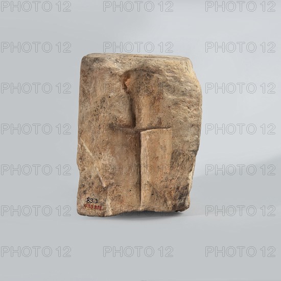 Hexagonal matrix for imprinting terracotta decorations, 1st–3rd century; Roman period, Tell Atrib, form; height 15.8 cm, width 15.3 cm, depth 12 cm, A roughly rectangular stone block with an uneven, weathered surface sits against a uniform pale gray background; the stone shows a carved, low-relief shape resembling a vertical pillar with a rounded top and a horizontal incision near its upper third, and the surface of the stone displays a range of earthy colors including tan, beige, brown, and hints of ochre and gray, with areas of darker mottling and some smoother lighter patches.
