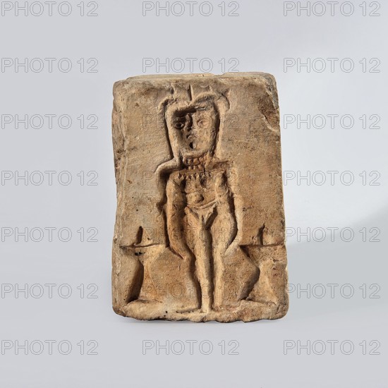 Hexagonal matrix for imprinting terracotta decorations, 1st–3rd century; Roman period, Tell Atrib; form; height 15.8 cm, width 15.3 cm, depth 12 cm, A rectangular tan-brown clay plaque with a shallow carved relief of a standing human-like figure centered on its front, the figure showing a head with stylized hair or headdress, facial features, a torso with suggestion of ornamentation around the neck and chest, and limbs that extend down to the base; the plaque surface is textured with small pits and rough areas and has worn edges and corners; a few symmetrical raised shapes flank the figure near the lower sides; the background around the plaque is a smooth pale gray with subtle gradient, and the visible colors include tan, brown, beige, gray, and a few darker brown spots.