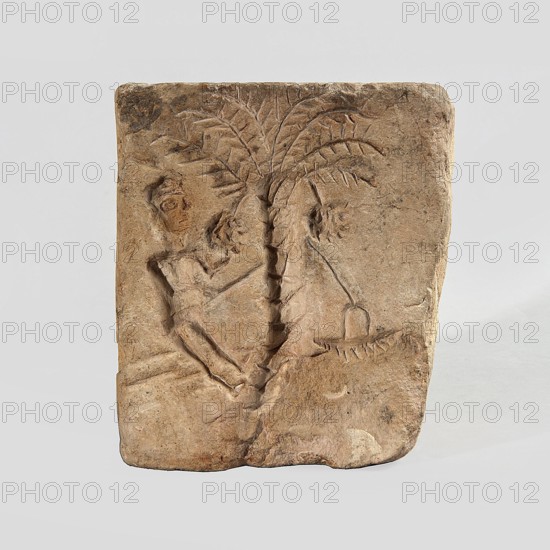 Hexagonal matrix for imprinting terracotta decorations, 1st–3rd century; Roman period, Tell Atrib, form; height 15.8 cm, width 15.3 cm, depth 12 cm, The image shows a rectangular carved stone tablet with a relief depicting a person reclining or leaning against a central palm-like tree with a textured trunk and radiating fronds; the person and tree are carved in shallow relief with visible tool marks and rough edges. The stone is mostly beige and light brown with some darker brown and grayish patches and worn areas; the background around the tablet is a plain light gray.