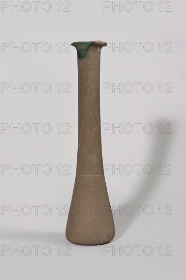 Bottle, 1st century BC–1st century, Egypt; vessel., A tall, narrow vase with a flared rim stands against a pale background. The vase has a textured surface in shades of brown and beige, becoming slightly darker toward the base. The rim shows a patch of green and a darker spot at the edge. The background is light gray to off-white, and a soft shadow in gray extends to the right of the vase.