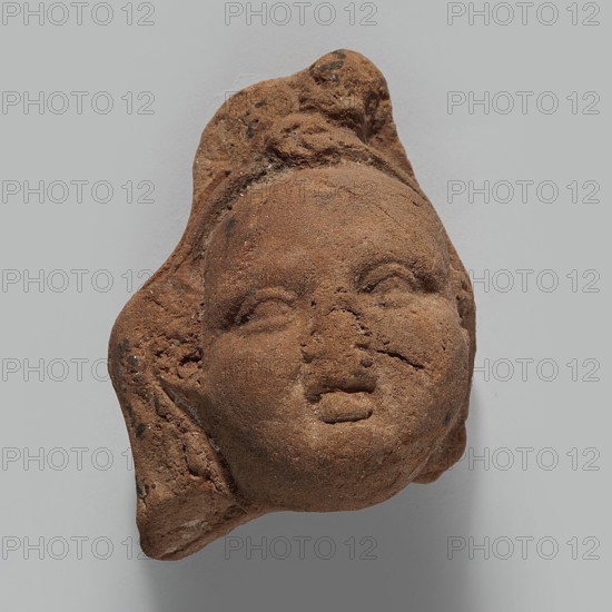 Head of Harpocrates; unknown creator; Harpocrates mythology; Hellenistic period, 332–30 BC, Egypt; figurine; height 5.4 cm, width 4 cm, depth 3.1 cm., A weathered clay or terracotta fragment shaped like a human head, with a rounded face, closed or partially closed eyes, a small nose, and slightly parted lips, showing rough, pitted texture and surface cracks; the piece has irregular edges and what appears to be a stylized hair or headdress form extending from the top and sides; colors visible include shades of brown, reddish-brown, and small darker brown or black flecks, all set against a neutral light gray background.