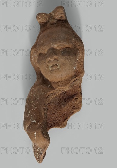 Harpocrates; preserved head and part of the right side of the figurine; traces of engobe on the entire surface and pink paint under the right elbow; Hellenistic or Roman period, Egypt; terracotta figurine; height 10.3 cm, width 3.8 cm, depth 3.4 cm., A fragment of a sculpted head and partial torso in a rough, brown-weathered material, showing a rounded face with simplified features and a small projecting mouth, a suggestion of closed eyes, and a smooth forehead topped by a small bowlike form; the surface has patches of lighter beige and small white speckles, with areas of darker brown and reddish-brown texture where the material is broken, all set against a neutral light gray background.