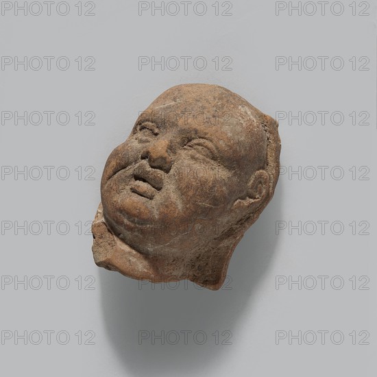 Harpocrates; fragment of a terracotta figurine head of Harpocrates; Hellenistic or Roman period, Egypt; figure; height 4.2 cm, width 3.2 cm, depth 2.3 cm; maker, inscription/author not specified., A sculpted head fragment carved from a brownish stone with surface variations in darker and lighter brown tones, showing a rounded face with closed or partially closed eyes, a flattened nose, and slightly parted lips; the features include chubby cheeks and a double chin, with a textured surface and some rough, chipped edges, especially at the base and around the back of the head, all set against a plain light gray background that casts a soft shadow beneath the fragment.