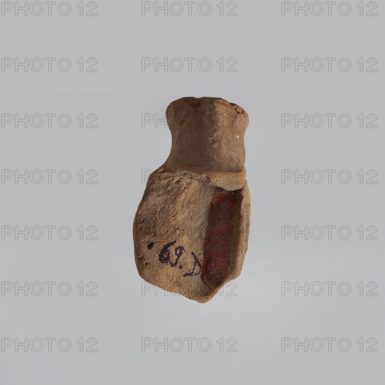 Serapis, fragment of a terracotta figurine head of Serapis with a kalathos on the head, lush curls and facial hair; terracotta; Hellenistic–Roman period, 323 BC–395 AD; Egypt; height 4.5 cm, width 2.6 cm, depth 2.5 cm;, A small irregularly shaped brown clay fragment with a rounded top and a flatter, rougher lower portion; the surface shows textured, worn areas and a recessed section on one side that reveals a darker brown-red interior; there are small dark specks and patches on the outer surface and subtle variations of light and shadow across the beige-gray background.