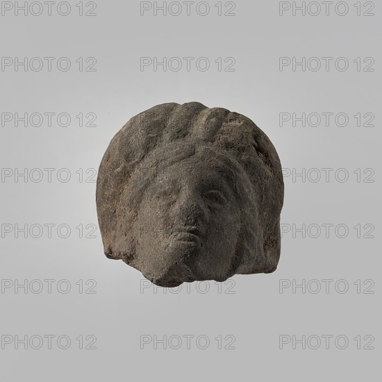Head of a female figurine, preserved from the chin up. High hairstyle with a wreath above the forehead, composed of oblique, wide strands of hair. Schematic facial features, wide nose. Unworked back, smooth, rounded. Dark gray clay. 332–30 BC; Hellenistic period, Egypt. Figurine. Height 3.3 cm, width 3.7 cm, depth 3.5 cm., A weathered stone sculpture fragment showing a human head with stylized hair and facial features, rendered in dark gray and brown tones with a rough, pitted texture, set against a plain light gray background.
