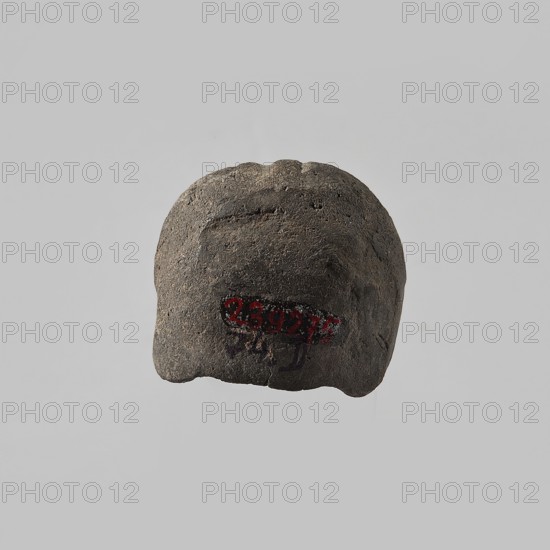 Head of a female figurine, preserved from the chin up. High hairstyle with a wreath above the forehead, composed of oblique, wide strands of hair. Schematic facial features, wide nose. Unworked back, smooth, rounded. Dark gray clay. 332–30 BC; Hellenistic period, Egypt. Figurine. Height 3.3 cm, width 3.7 cm, depth 3.5 cm., A roughly rectangular, rounded object with an uneven, textured surface sits against a plain light gray background; the object is primarily dark gray to charcoal with areas of lighter gray and a few small spots of darker shadow, and there is a small area of bright red pigment near the lower center of the object.