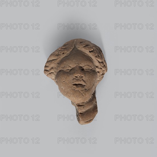 Head of a terracotta female figurine with part of the neck and a fragment of a robe preserved. Hairstyle parted in the middle. Facial features quite simplified and rounded. Light brown clay with uneven firing — center of the gray color break. Dated 332–30 BC, Hellenistic period, Egypt. Figurine; height 3.8 cm, width 3 cm, depth 2.1 cm., A weathered brown stone fragment shaped like a human face with stylized hair and softened facial features casting a faint shadow on a pale gray background, showing textured rough surfaces and areas of erosion with visible contours of a nose, closed mouth, and suggestion of eyes.