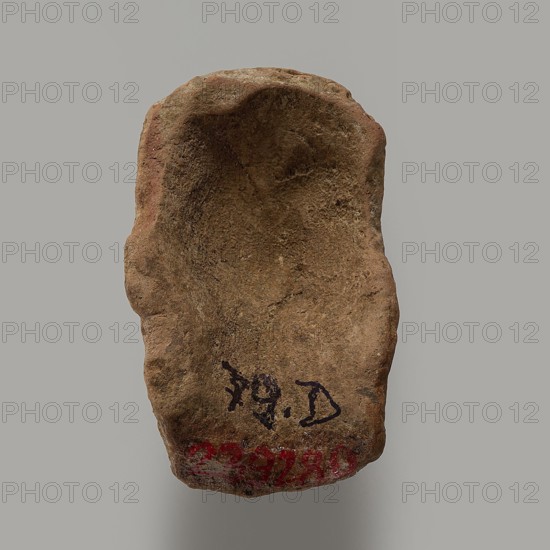Fragment of a figurine - head of a man. Head of a male figurine. High forehead, above it fragments of short hair; large, wide-open eyes with distinct upper and lower eyelids; slanting, long eyebrows above the eyes; large nose, small mouth, longish ears, neck with a distinct apple. Pink clay, light beige engobe. 332–30 BC; Hellenistic period, Egypt. Figurine; height 5 cm, width 3.2 cm, depth 2.3 cm., A roughly shaped, irregular brown stone with a hollowed, concave upper surface and a textured, grainy appearance; the stone shows areas of darker brown shading and small rough patches, with some reddish-brown tones along one edge and hints of gray in the background.