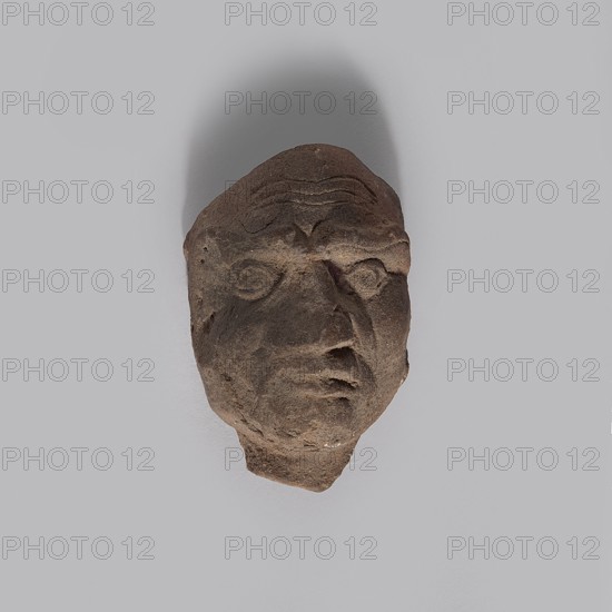 Head of a man. Fragment of a terracotta figurine showing the head of an elderly man. Thin face with a bald skull; three long horizontal wrinkles on the forehead; wide eyebrows connecting above the nose forming two vertical wrinkles in a V-shape; sharp eyes with distinct upper and lower eyelashes, irises, and dark pupils. Crooked, rather large nose; closed mouth; vertical furrow on the chin; narrow neck. Pinkish, gray-burnt clay. Hellenistic period, Egypt, 332–30 BC. Figurine; height 4 cm, width 2.8 cm, depth 1.7 cm., A roughly carved stone head with a rough texture and irregular shape sits against a smooth pale gray background, the stone showing mostly brown and dark brown tones with some lighter tan areas; the face has deeply incised features including prominent brow ridges, hollowed eye sockets, a pronounced nose, and a downturned mouth, with visible tool marks and surface pitting, and a shadow falls above the object creating a soft darker gray arc on the background.