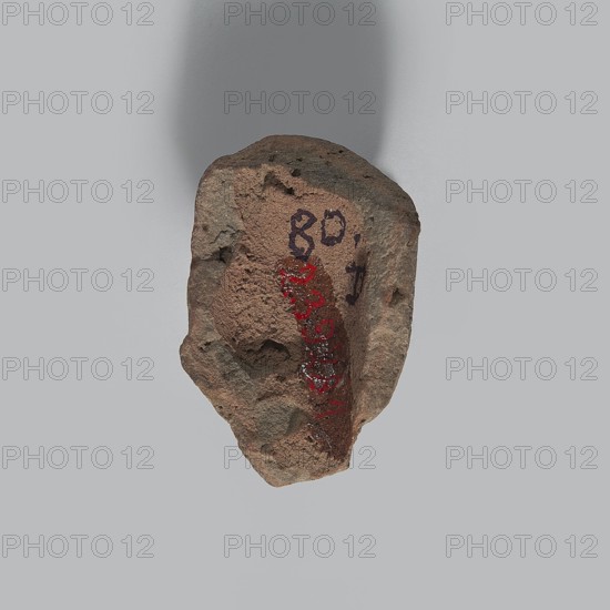 Head of a man. Fragment of a terracotta figurine showing the head of an elderly man. Thin face with a bald skull; on the forehead three very long horizontal wrinkles; wide eyebrows connecting above the nose forming two vertical wrinkles that meet in a V-shape; sharp eyes with distinct upper and lower eyelashes, irises, and dark pupils. Crooked, rather large nose; closed mouth; vertical furrow on the chin; narrow neck. Pinkish, gray-burnt clay. Hellenistic period, Egypt, 332–30 BC. Figurine; height 4 cm, width 2.8 cm, depth 1.7 cm., A rough, irregularly shaped rock with a textured surface in shades of brown and gray sits against a pale gray background, showing a few small pits and crevices and an area with a darker reddish-brown patch; there are also traces of darker gray markings and a vertical streak of red on one face.