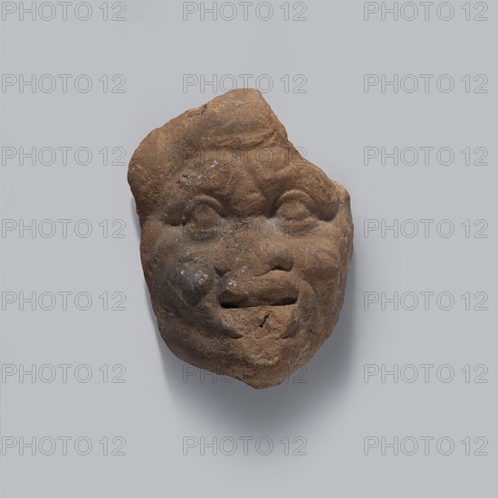 Head of a man. Fragment of a terracotta figurine of a man in a caricatured style. The face is wide with a low forehead with wrinkles and small eyes with prominent upper eyelashes. The nose is wide and flat with protruding cheeks. The face has a large, open, thick mouth. Pinkish-brown clay, fired. 332–30 BC; Hellenistic period, Egypt. Figurine. Height 4 cm, width 2.8 cm, depth 1.7 cm., A weathered brown stone fragment shaped like a human face with a rounded forehead, pronounced brows, closed eyes, a broad nose, and a slightly open mouth, showing some surface pitting and lighter spots, set against a smooth pale gray background with a soft shadow beneath; visible colors include brown, pale gray, and small areas of off-white.