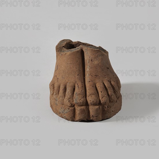 Fragment of a terracotta figurine showing two feet placed side by side on a flat, rounded base., Terracotta. Hellenistic or Roman period, Egypt, 323 BC–476 AD. Figurine. Height 4 cm, width 5 cm, depth 6.3 cm. ., A small clay sculpture of a pair of feet sits on a smooth pale gray surface against a pale gray background; the sculpture is brown with darker brown and gray speckling, showing toes and worn, cracked texture with rough edges and uneven surfaces, casting a soft shadow to one side.