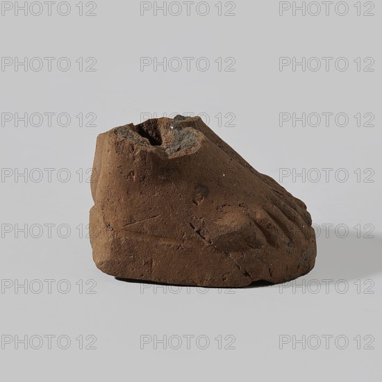 Fragment of a terracotta figurine showing two feet placed side by side on a flat, rounded base. Hellenistic or Roman period, Egypt, 323 BC–476. Figurine; height 4 cm, width 5 cm, depth 6.3 cm., A roughly foot-shaped object made of brown, earthy material sits on a light gray surface against a pale gray background; the object has a textured, uneven surface with a few small dark gray and black patches and some tiny lighter specks, visible depressions resembling toes and a hollowed top, and casts a soft shadow beneath and slightly to the right.