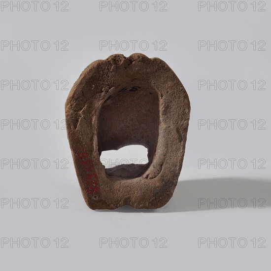 Fragment of a terracotta figurine showing two feet placed side by side on a flat, rounded base. Hellenistic or Roman period, Egypt, 323 BC–476 AD. Figurine; height 4 cm, width 5 cm, depth 6.3 cm., A weathered brown hollow stone with an irregular oval opening sits upright on a pale gray surface against a light gray background, showing rough textured surfaces, rounded edges, and small pits and indentations, with a shadow cast to the right and a few darker brown variations across the stone.