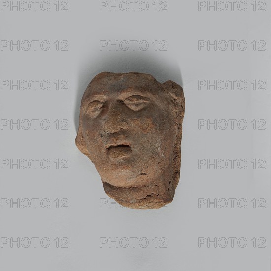 Fragment of the head of a terracotta female figurine. Fragmentarily preserved head depicting a woman: face and upper part of the neck. Narrow, open mouth with wide lips; large eyelids giving the eyes a half-closed appearance; fairly wide nose. Hairstyle unpreserved. Made of fairly poor quality clay with minor cracks especially on the neck and chin and visible grains; surface slightly effaced. Hellenistic or Roman period, Egypt, 323 BC–476 AD. Figurine; height 3.6 cm, width 3.0 cm, depth 2.3 cm., A worn sculpted head fragment in shades of brown and reddish-brown rests against a light gray background; the fragment shows an abstracted human face with closed or partially closed eyes, a nose, and a slightly open mouth, and the surface displays rough, pitted texture with areas of darker and lighter brown.