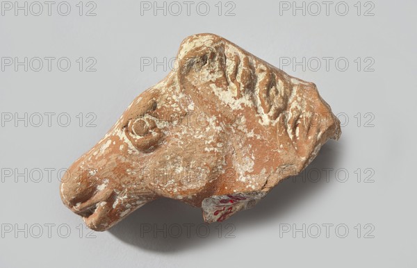 Head of a terracotta horse figurine. Terracotta head with part of the neck and mane of a horse. The animal's eyes and muzzle are marked. The mane, depicted in a long strip, falls to the right side of the head. The surface is heavily worn, with partially erased convex details. The darker original clay surface and remnants of red paint, are preserved in the recesses. Dated 332–30 BC; Hellenistic period, Greater Greece. Figurine. Height 3.3 cm, width 5.3 cm, depth 2.2 cm., A worn, irregularly shaped terracotta fragment resembling an animal head with textured surface and weathered edges, showing a few raised rounded features where an eye and snout appear, along with shallow carved indentations suggesting fur or mane detail; the object displays a mix of reddish-brown clay and patches of lighter beige or off-white encrustation, and it rests on a smooth light gray background casting a soft shadow beneath.