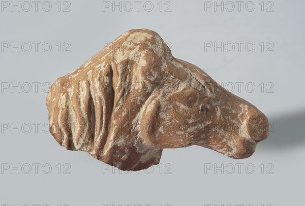 Head of a terracotta horse figurine. Terracotta. Head with part of the neck and mane of a horse; the animal's eyes and muzzle are marked. The mane, depicted in a long strip, falls to the right side of the head. The surface is heavily worn, with partially erased convex details. The darker original clay surface and remnants of red paint, are preserved in the recesses. Hellenistic period, Greater Greece, 332–30 BC. Figurine. Height 3.3 cm; width 5.3 cm; depth 2.2 cm., A weathered, irregularly shaped stone with a rough surface showing a mix of brown and tan areas and lighter beige patches, featuring deep grooves and rounded protrusions that suggest layered textures; the stone rests against a smooth, pale gray background with a soft shadow to one side.