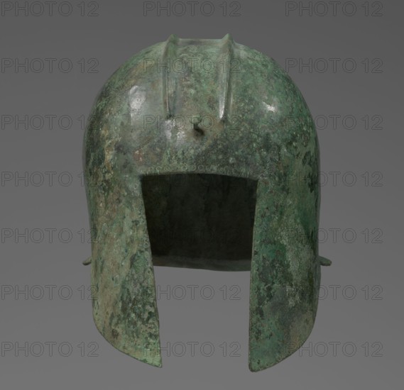 Bronze helmet with rectilinear cutouts for the eyes, nose, mouth, and chin, with two prominent reinforcement ribs. 550–500 BC, Italy, armament. Height 26 cm, width 19.3 cm, depth 25 cm, weight 0.994 kg., A single metal helmet is shown from the front, with a rounded dome and two downward flaring cheek guards creating a large rectangular face opening; the surface is uneven and pitted with areas of rough texture and small raised ridges along the crown, and there are a few small protruding studs on the top and sides. The visible colors are shades of green and greenish-gray with patches of darker green, muted brown, and some lighter greenish highlights, set against a plain neutral gray background.