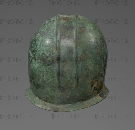 Bronze helmet with rectilinear cutouts for the eyes, nose, mouth, and chin and two prominent reinforcements ribs. 550–500 BC, Italy, armament. Height 26 cm, width 19.3 cm, depth 25 cm, weight 0.994 kg., A rounded helmet-like object with a domed top and a short brim shown against a plain gray background, displaying a mottled surface in shades of green, teal, dark green, brown, and rust, with a pair of raised ridges running from the top toward the brim and areas of darker and lighter patina and spotting across its surface.