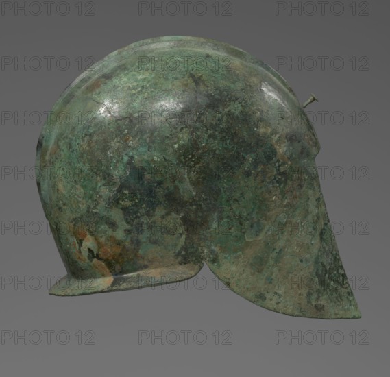 Bronze helmet with rectilinear cutouts for the eyes, nose, mouth, and chin; two prominent reinforcement ribs. 550–500 BC; Italy . Armament. Height 26 cm, width 19.3 cm, depth 25 cm, weight 0.994 kg., A single metallic object with a rounded dome and an extended rear flap, showing a textured surface with patina in shades of green and darker green, along with areas of brown, rust, and muted gray; the object has a small protruding peg near the top rear and a narrow brim at the front edge, all set against a plain gray background.