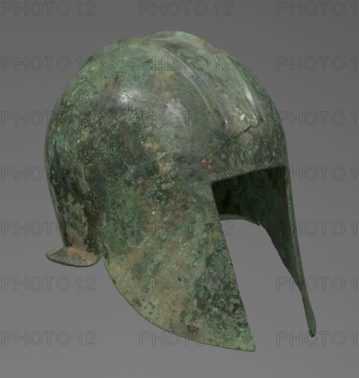 Bronze helmet with rectilinear cutouts for the eyes, nose, mouth, and chin. The helmet has two prominent reinforcements ribs. 550–500 BC, Italy, armament. Height 26 cm, width 19.3 cm, depth 25 cm, weight 0.994 kg., The image shows an aged metal helmet with a rounded crown and a forward-projecting nose guard, displaying a weathered, mottled surface in shades of green, turquoise, brown, and dark gray, with patches of lighter and darker discoloration and areas of subtle sheen; the helmet has an extended cheek and neck guard and a slight raised ridge along the top, all set against a plain medium-gray background.