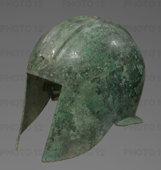 Bronze helmet with rectilinear cutouts for the eyes, nose, mouth, and chin; two prominent reinforcements ribs. 550–500 BC, Italy, armament. Height 26 cm, width 19.3 cm, depth 25 cm, weight 0.994 kg., A metal helmet with a rounded dome and extended cheek guards is shown against a plain gray background, exhibiting a mottled surface of green and darker green patina with areas of brown and gray, an uneven texture with spots of lighter green and pale gray, a raised central ridge on the dome, small rounded rivet-like bumps along the lower edge, and a small flared projection at the rear, with the interior visible as darker metal tones and hints of the exterior patina.