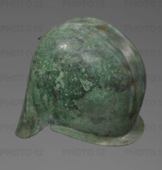 Bronze helmet with rectilinear cutouts for the eyes, nose, mouth, and chin; the helmet has two prominent reinforcing ribs. 550–500 BC, Italy, armament; height 26 cm, width 19.3 cm, depth 25 cm, weight 0.994 kg., An aged metal helmet shown from a side angle with a rounded dome and a small projecting brim, exhibiting a mottled surface of green patina with areas of darker green, lighter green, and patches of brown and gray; the background is a uniform gray.