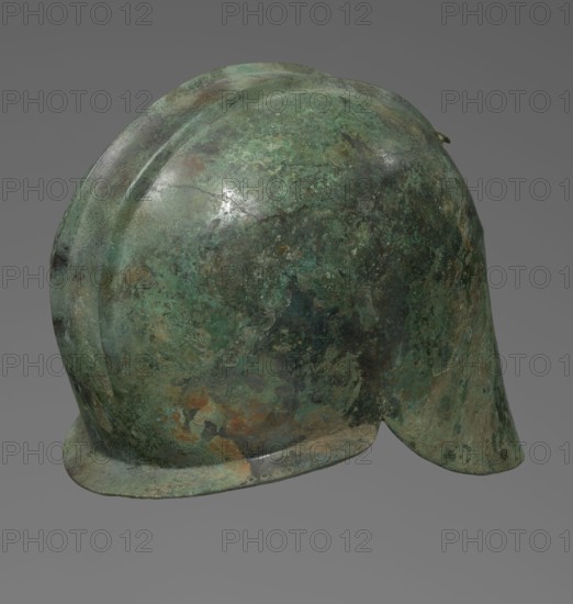 Bronze helmet with rectilinear cutouts for the eyes, nose, mouth, and chin; two prominent reinforcements ribs. 550–500 BC, Italy ; armament. Height 26 cm, width 19.3 cm, depth 25 cm, weight 0.994 kg., A weathered helmet with a rounded dome and a flared back, showing a patina of green and turquoise with areas of darker green, brown, and small patches of orange-brown corrosion; the surface has mottled textures and subtle shiny highlights, and the helmet sits against a plain gray background.