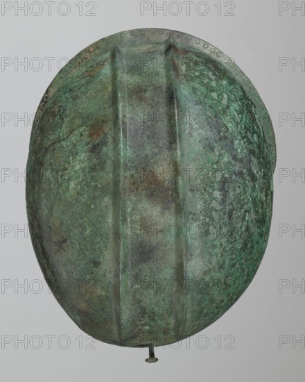 Bronze helmet with rectilinear cutouts for the eyes, nose, mouth, and chin; the helmet has two prominent reinforcements ribs. 550–500 BC; Italy . Armament. Height 26 cm, width 19.3 cm, depth 25 cm, weight 0.994 kg., An oval, helmet-like object with a textured, mottled surface showing shades of green, turquoise, brown, and gray, featuring three raised ridges running lengthwise, a rounded rim with a row of small circular impressions along its edge, and a short metal peg protruding from the bottom; the background is a smooth pale gray.
