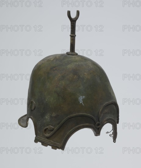 Bronze helmet, 4th–3rd century BC, Italy, armament; height 27 cm without cheek guards, width 19.8 cm, depth 24 cm, weight 0.738 kg without cheek guards, A worn metallic helmet with a rounded dome and an upright forked finial on top, showing a mottled surface in shades of green, brown, dark bronze, and hints of black, with a small pale bluish spot; the lower edge features several decorative curved flanges and scroll-like elements in similar dark bronze and brown tones, and there are a few small hooks and looped attachments along the rim.
