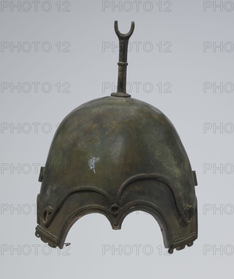 Bronze helmet, 4th–3rd century BC, Italy, armament; materials: bronze; height 27 cm without cheek guards, width 19.8 cm, depth 24 cm, weight 0.738 kg without cheek guards., The image shows a metal helmet with a rounded dome and a small vertical protrusion on top ending in a forked shape; the surface of the helmet displays a mottled patina in shades of green, brown, and dark gray with a small irregular lighter spot. The lower edge of the helmet features a scalloped outline with decorative raised ridges and curved metal bands, and there are small hooked or looped metal fittings at the bottom edges. The background is a smooth, light gray.