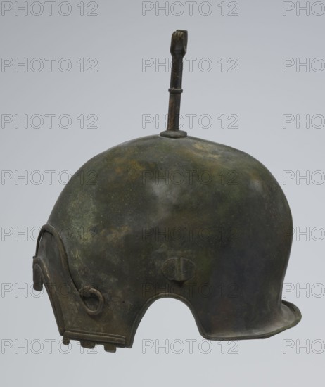 Bronze helmet, 4th–3rd century BC, Italy, armament; height 27 cm without cheek guards, width 19.8 cm, depth 24 cm, weight 0.738 kg without cheek guards., A worn metal helmet viewed from the side with a rounded dome and a vertical fin-like projection rising from the top; the surface shows mottled patches of dark green, brown, and muted gold with areas of darker gray and black; a curved ear-cover piece with a looped detail and a small rectangular segmented attachment is visible toward the lower front, and a rounded cutaway near the base forms an arch; the rim along the lower edge is slightly flared and uneven in color, and the vertical projection has a darker brown tone with a small ring-like band about its shaft.
