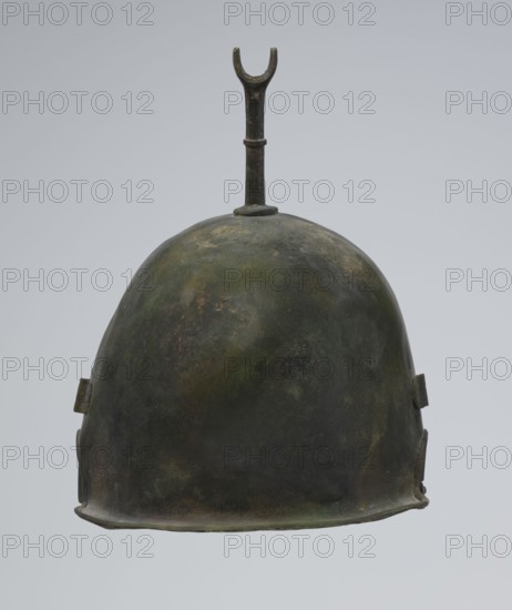 Bronze helmet, 4th–3rd century BC, Italy, armament; height 27 cm without cheek guards, width 19.8 cm, depth 24 cm, weight 0.738 kg without cheek guards, A single metal helmet with a rounded dome shape and a short cylindrical stem topped by a U-shaped prong extending upward, showing a mottled surface with dark green, olive, brown, and hints of black and gray tones; the edges are slightly irregular and a few small rectangular tabs project from the sides, and the background is a smooth pale gray.