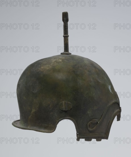 Bronze helmet, 4th–3rd century BC, Italy, armament; height 27 cm without cheek guards, width 19.8 cm, depth 24 cm, weight 0.738 kg without cheek guards., A rounded metal helmet with a short brim and a cutaway section at the lower edge, shown in profile, with a vertical rod fixed at the top; the surface shows a mottled patina in shades of green, dark green, brown, and hints of black, with areas of lighter greenish and yellowish discoloration, and a decorative raised ridge and curved motif near the rear edge.