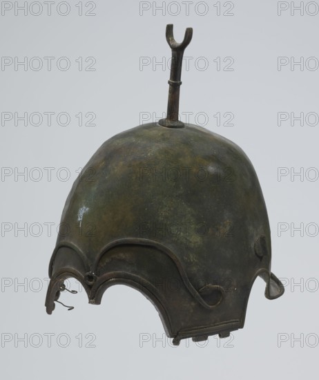 Bronze helmet, 4th–3rd century BC, Italy, armament; height 27 cm without cheek guards, width 19.8 cm, depth 24 cm, weight 0.738 kg without cheek guards, A bronze helmet with a rounded dome showing a mottled surface of green, dark green, brown, and hints of black, topped by a vertical U-shaped finial on a short stem; the lower edge of the helmet features scalloped arches and raised decorative ridges with curling end details, and a few small looped metal hooks hang from one of the arches.