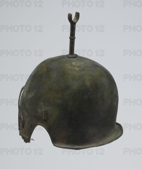 Bronze helmet, 4th–3rd century BC, Italy, armament; height 27 cm without cheek guards, width 19.8 cm, depth 24 cm, weight 0.738 kg without cheek guards, A metal helmet with a rounded dome shape and a short brim along the lower edge, showing a mottled surface in dark green, brown, and black tones, with areas of lighter green and muted yellowish discoloration; a vertical metal rod topped with a forked or yoke-like piece protrudes from the top center; the helmet has a cutaway around the lower front edge and a small circular projection on the side, with visible seams and slight irregularities in texture and patina.