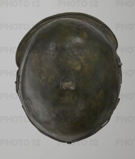 Bronze helmet, 4th–3rd century BC, Italy, armament; height 27 cm without cheek guards, width 19.8 cm, depth 24 cm, weight 0.738 kg without cheek guards, A close-up view of a rounded metallic object with an uneven, slightly dented surface showing a mix of dark green, olive, brown, and hints of black tones, with a worn central protrusion and a rim that curves around the top edge, slight darker shadows around the sides, and a neutral light gray background.