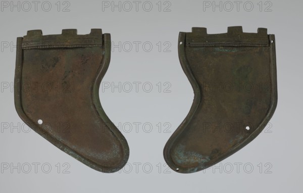 Bronze helmet, 4th–3rd century BC, Italy, armament; height 27 cm without cheek guards, width 19.8 cm, depth 24 cm, weight 0.738 kg without cheek guards., Two curved metal plates with irregular rounded shapes lie side by side against a neutral light gray background, each showing a patina of dark brown and green with areas of bluish-green corrosion. The left plate has a raised rim and a few small darker spots and streaks across its surface, along with a small circular hole near its lower edge, while the right plate mirrors the shape with similar raised edges, scattered darker patches, and a small hole near its lower outer edge; both pieces have a slightly rough, mottled texture and a thin raised strip near their top edges.