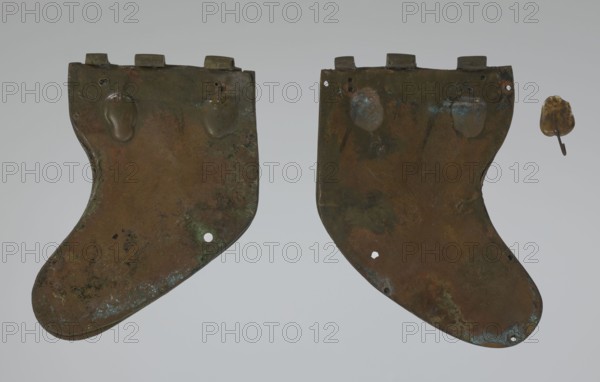 Bronze helmet, 4th–3rd century BC, Italy, armament; height 27 cm without cheek guards, width 19.8 cm, depth 24 cm, weight 0.738 kg without cheek guards, A pair of large, irregularly shaped metal plates with rounded lower edges are shown against a gray background, each plate displaying patinated surfaces in shades of brown, green, and blue with darker mottled areas; the left plate has a few raised oval bumps near its top and a small hole near its lower edge, while the right plate mirrors the left in shape with similar raised areas and small holes and shows patches of bluish-green corrosion; to the right of the pair floats a small, separate metal fragment that is golden-brown with a short protruding piece.