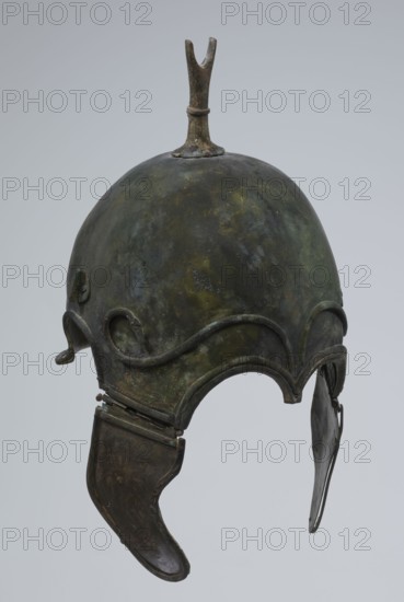 Bronze helmet, 4th–3rd century BC, Italy, armament; height 34 cm, width 20.2 cm, depth 22.5 cm, weight 1.035 kg., A metal helmet with a rounded dome and a short vertical fin on top, showing a weathered surface with mottled green, brown, and dark gray patina, featuring decorative raised curved ridges around the lower dome and hinged cheek guards that are darker brown with worn edges, all set against a pale gray background.