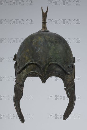 Bronze helmet, 4th–3rd century BC, Italy, armament; height 34 cm, width 20.2 cm, depth 22.5 cm, weight 1.035 kg, An aged metal helmet with a rounded dome and a vertical fin at the top, showing mottled patches of green, dark green, brown, and hints of bluish tones; the front edge has a decorative wavy ridge forming two rounded arches above the eye openings, and two elongated cheek guards hang down on each side with similar mottled green and brown patina.