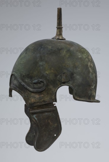 Bronze helmet, 4th–3rd century BC, possibly Italy; armament; height 34 cm, width 20.2 cm, depth 22.5 cm, weight 1.035 kg., A metal helmet with a rounded dome and a short vertical spike on top, showing a mottled surface in shades of dark green, brown, and hints of gray, with a curved decorative ridge along the side and a large cutout for the ear; a hinged cheek guard hangs below with a worn surface in dark green and brown tones and traces of lighter green patina, all set against a pale gray background.
